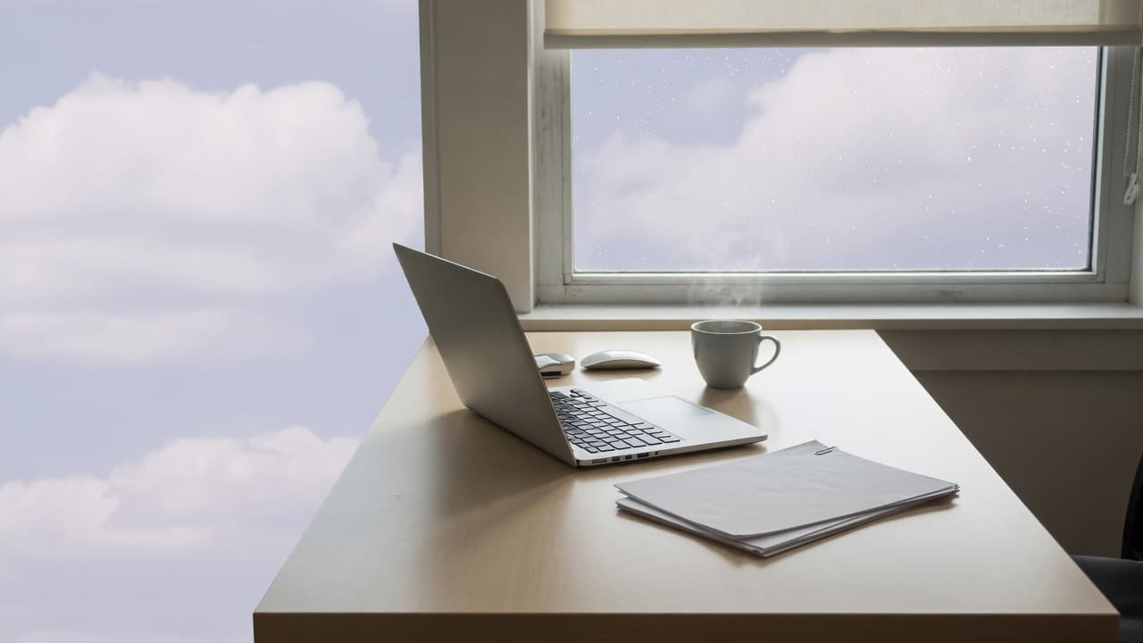A quiet desk with a laptop, a mug, and a short stack of paper by a window.